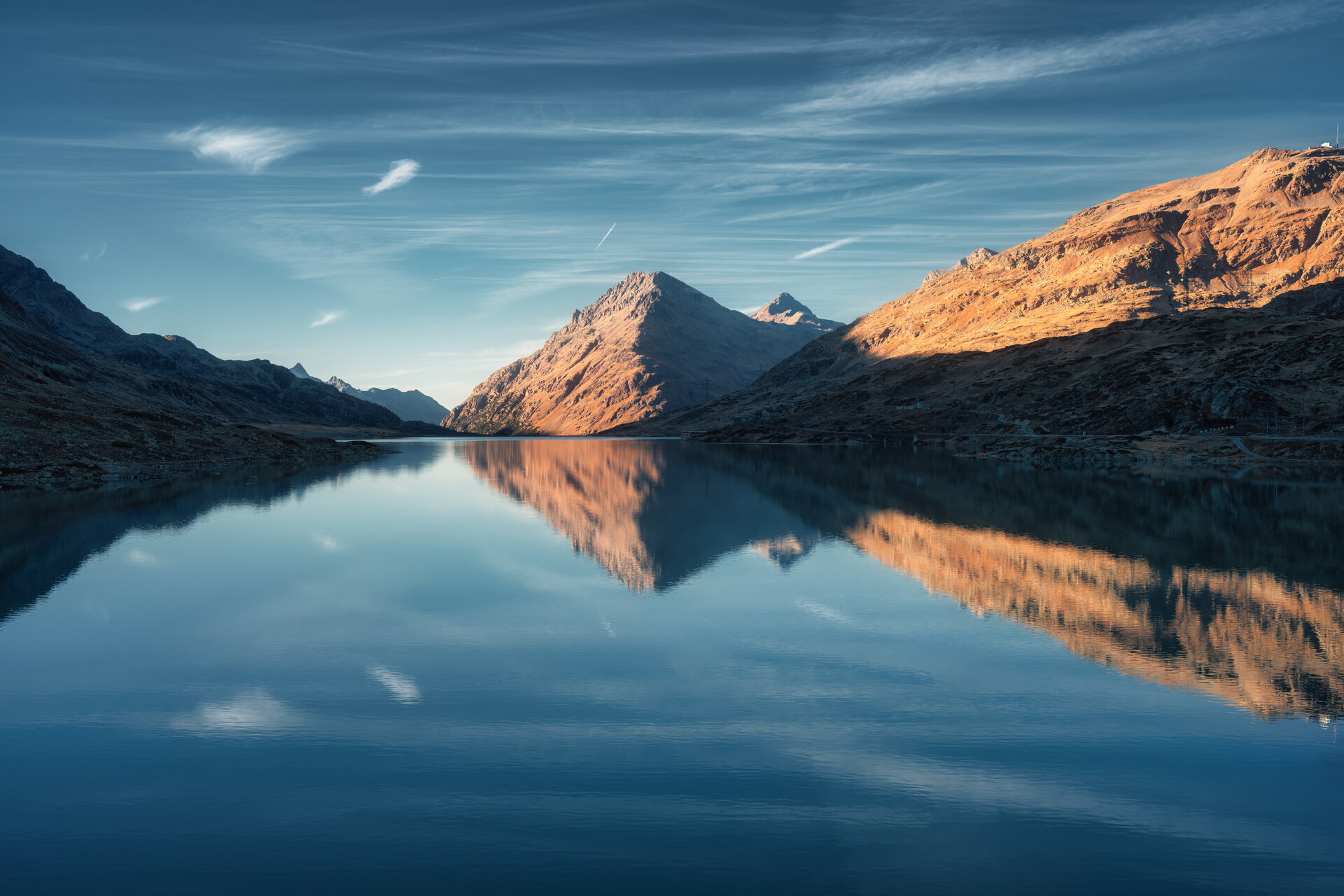 Sunset casts a serene glow on mountain peaks mirrored in a tranquil alpine lake in autumn. Lake Bianco, Switzerland in fall. Landscape with mountains, reflection in water, blue sky. Picturesque scene